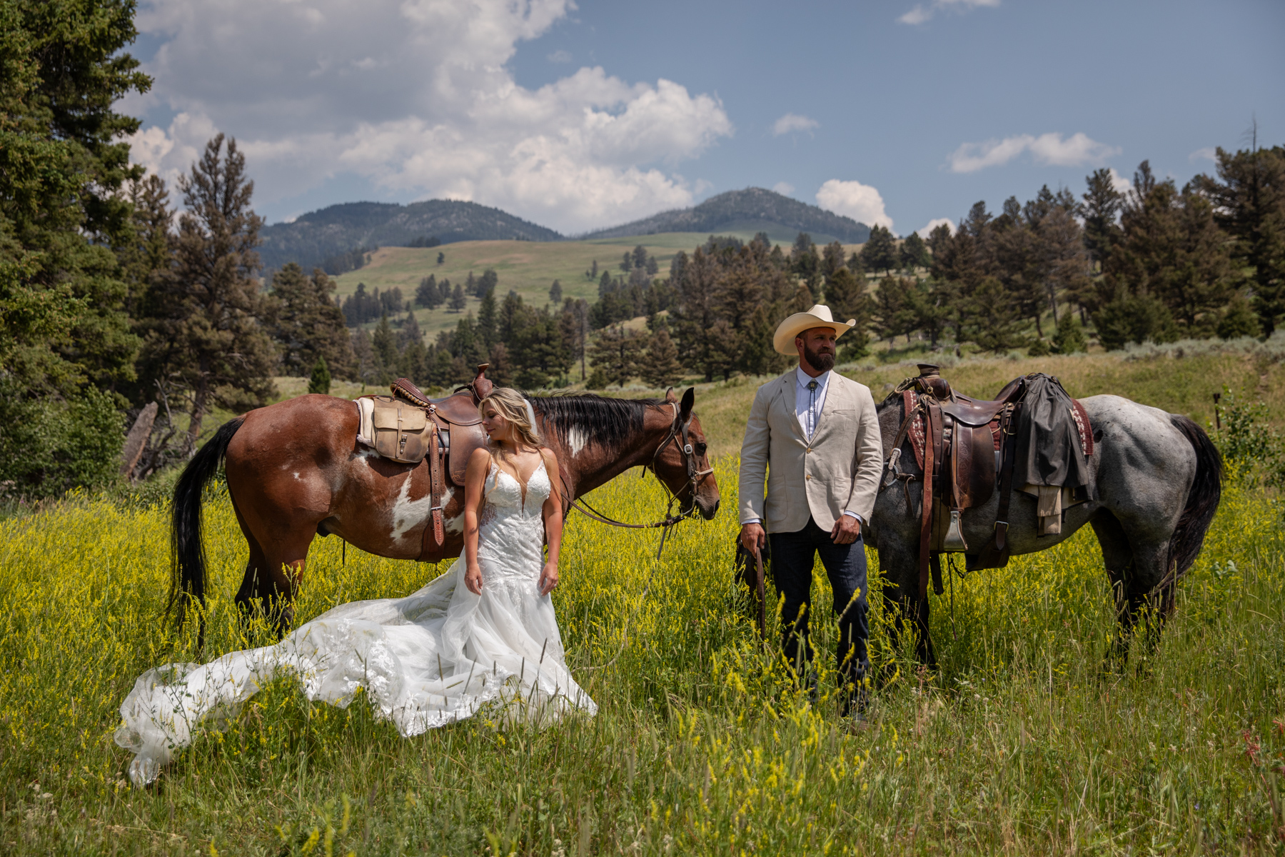 Bride and groom pose in from of their horses in a beautiful field of yellow flowers and green mountains in the background. 