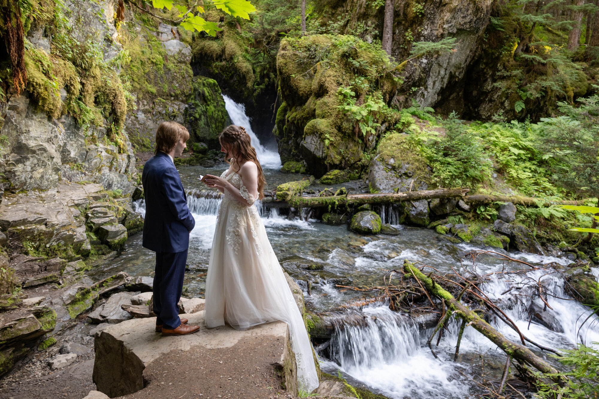 Couple standing beside a forest waterfall in Girdwood, Alaska during an intimate elopement surrounded by lush greenery.