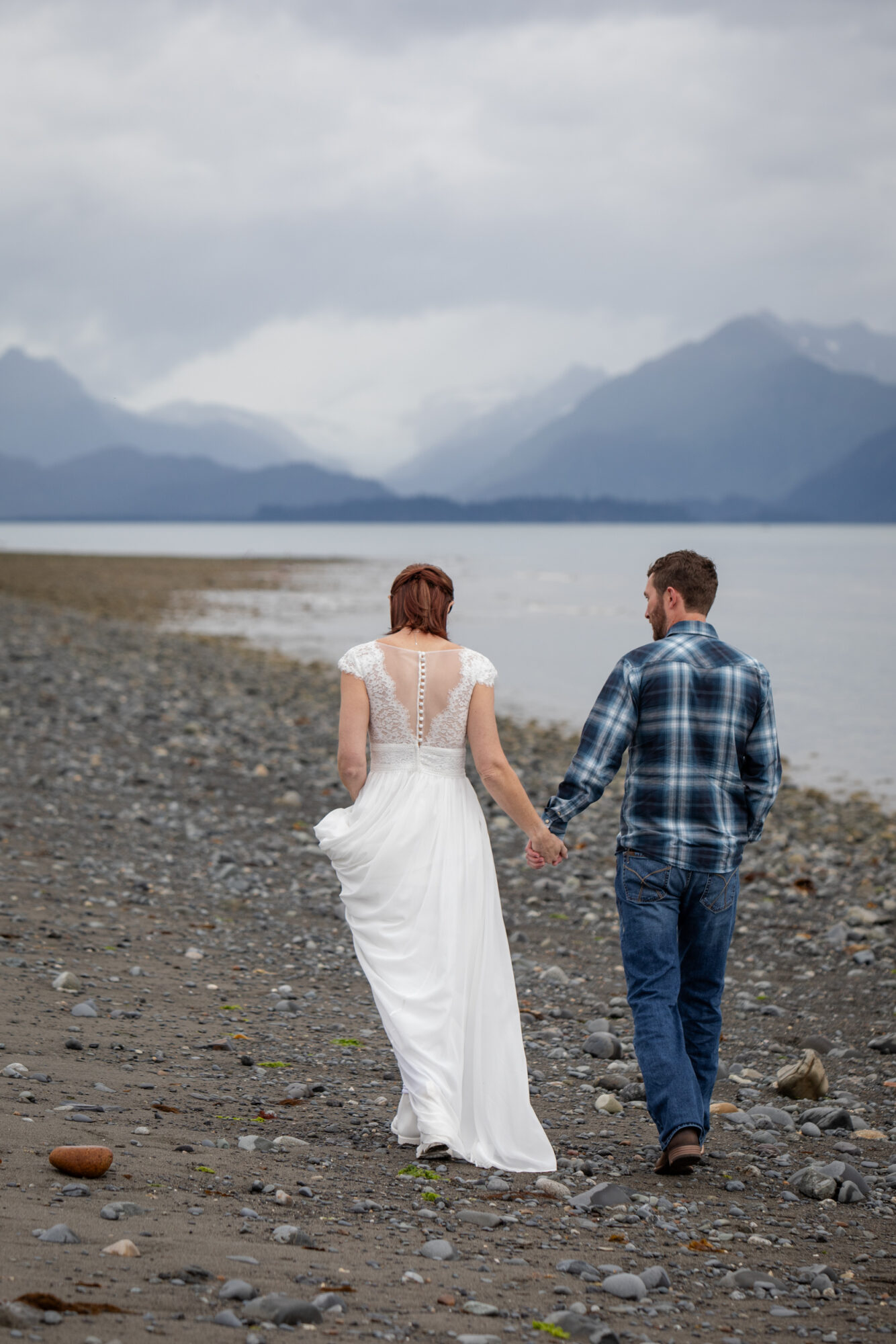 couple walking along Bishop beach during their Homer Alaska elopement with ocean and layered mountains in the distance