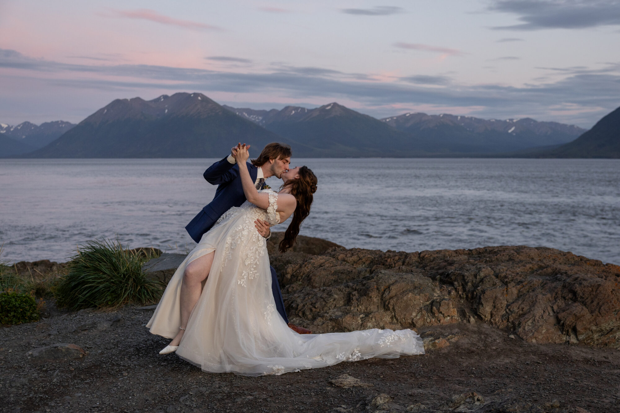Couple embracing along a rocky shoreline near Girdwood, Alaska with ocean views and mountains at sunset during an elopement.