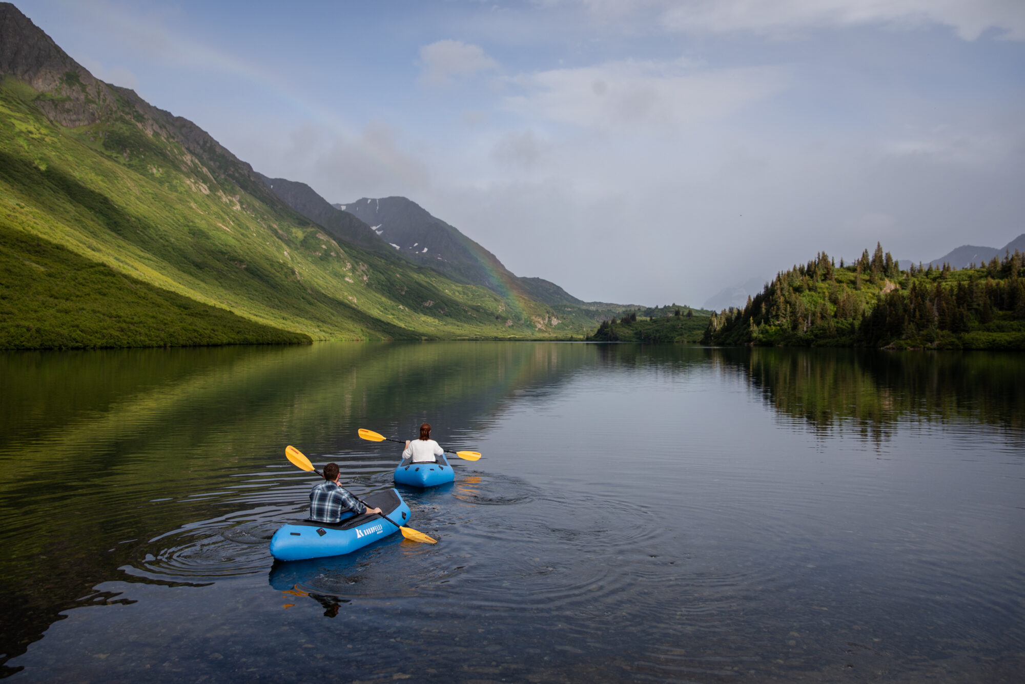 couple kayaking across a calm alpine lake in Kachemak Bay State Park during their Homer Alaska elopement with a rainbow over the mountains
