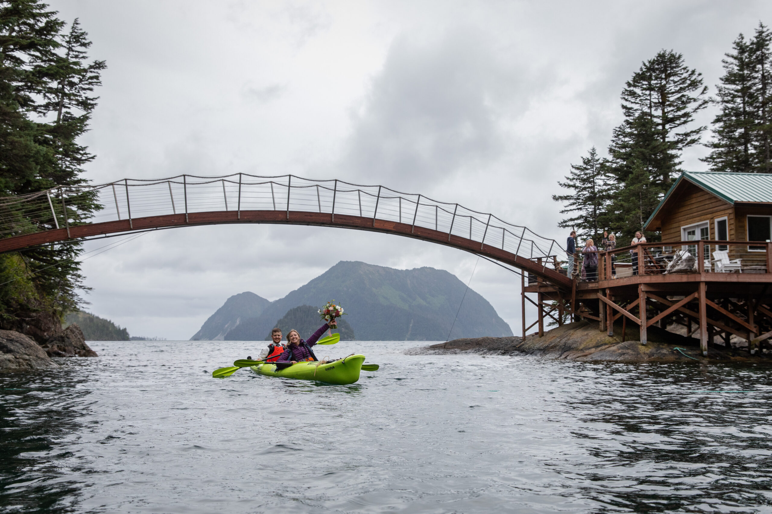 A bride and groom paddle in a kayak under a bridge after they figured out how to elope.