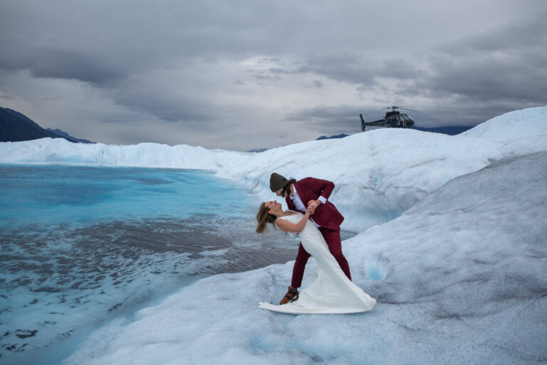 Grooms dips bride by a blue glacier pool in Alaska while their helicopter waits for them.
