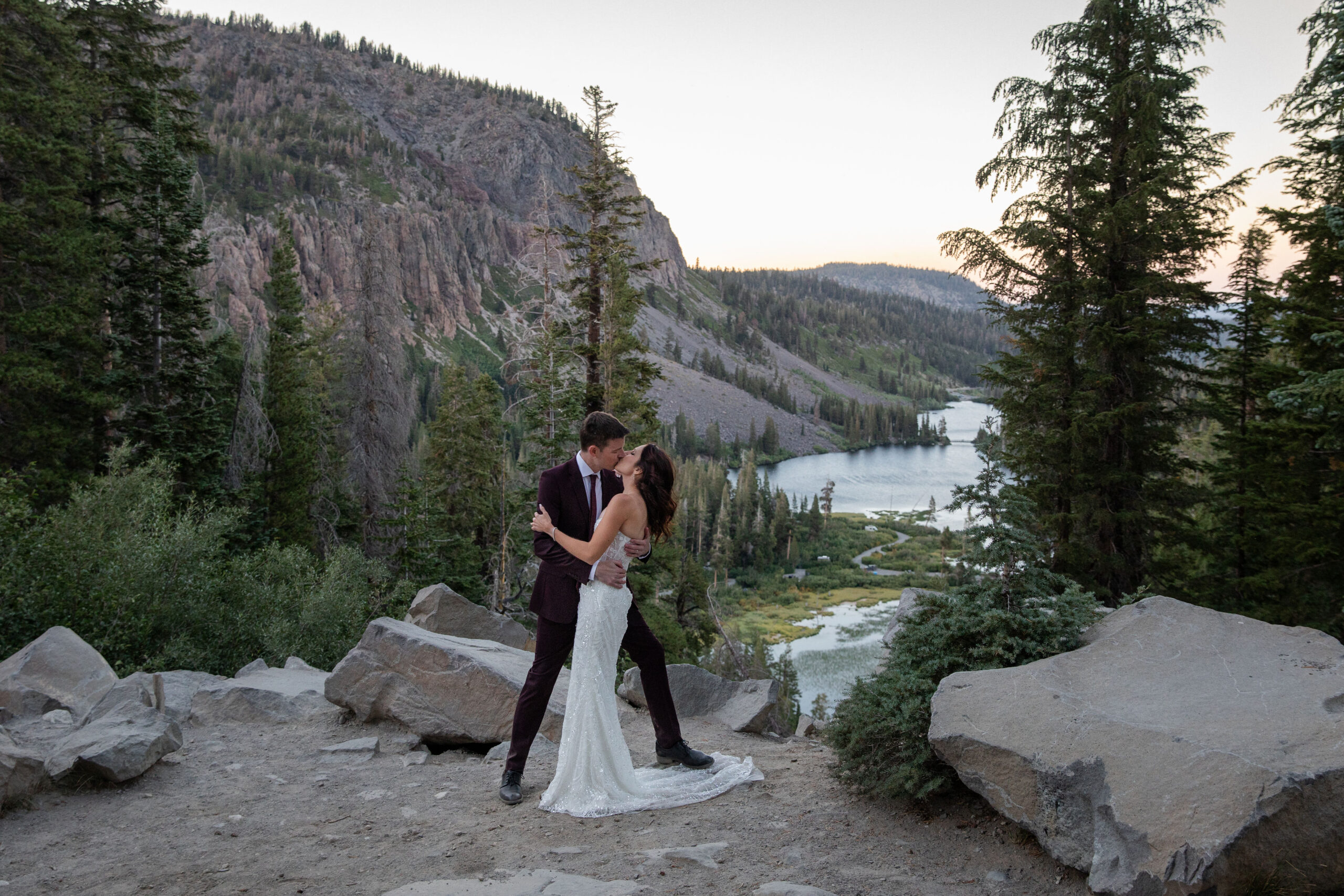 A bride and groom share an intimate kiss on a rocky overlook surrounded by towering pine trees and a serene mountain lake in the distance. The groom wears a deep burgundy suit, while the bride stuns in a white lace wedding dress. The backdrop features rugged cliffs and a tranquil alpine setting, bathed in soft natural light.