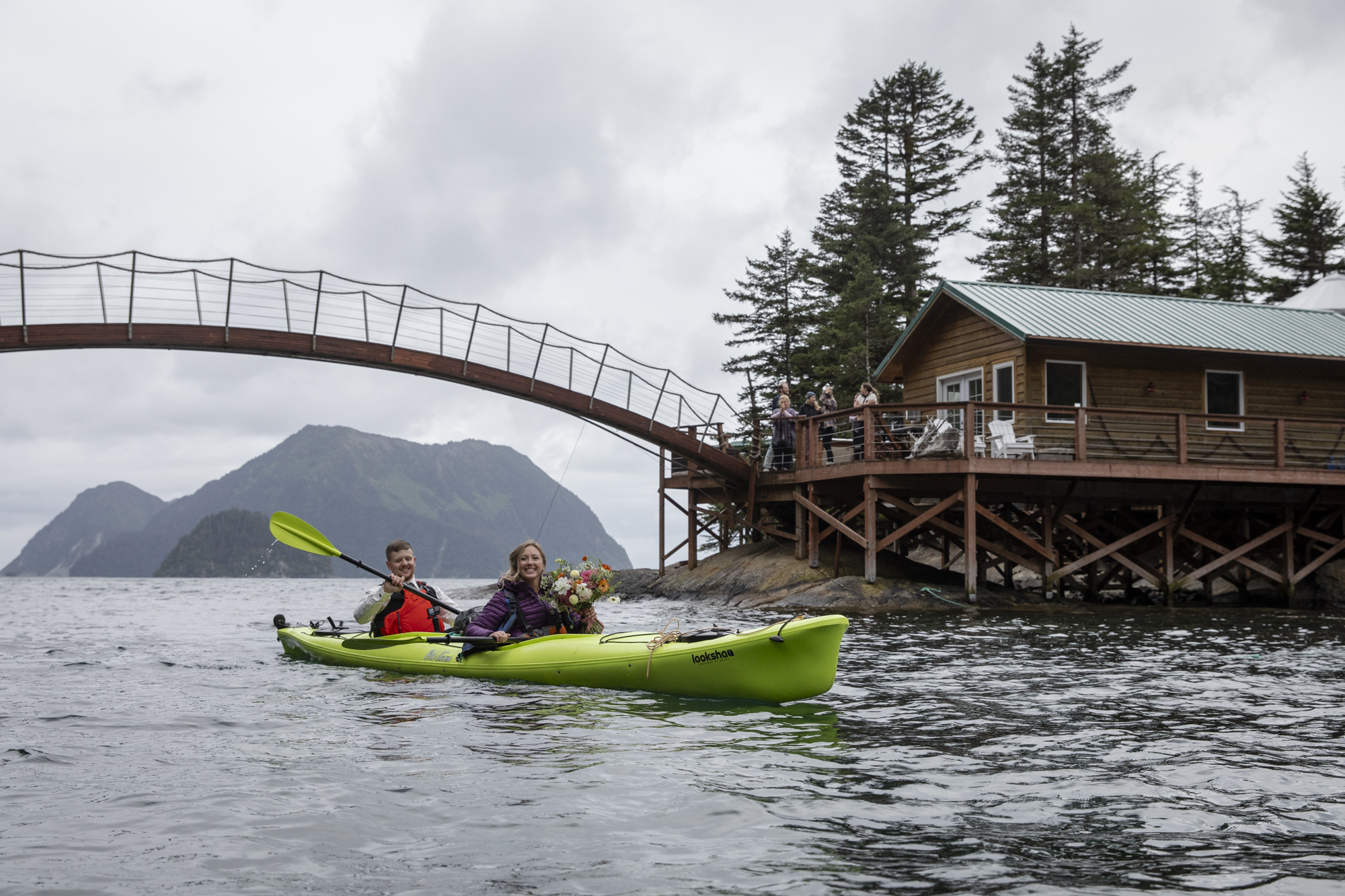 Bride and groom paddle a sea kayak on the way back to the lodge as guests cheer from the shoreline. 