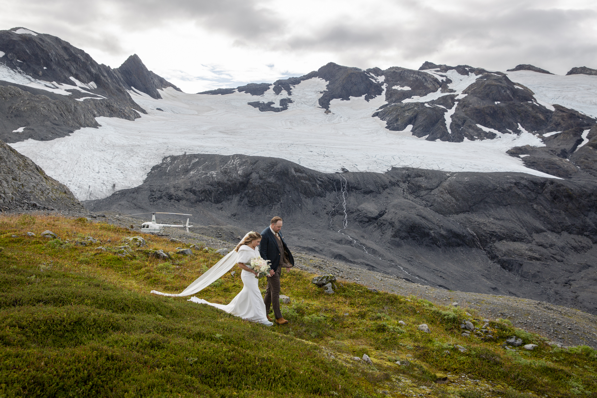 Couple walks down a green hillside with their helicopter waiting in the background in front of towering Alaskan peaks.  Helicopters  earn a solid spot as one of our adventure elopement ideas!