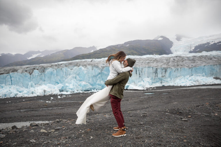 A groom lifts his bride in the air as their noses touch in front of a glacier in Alaska.