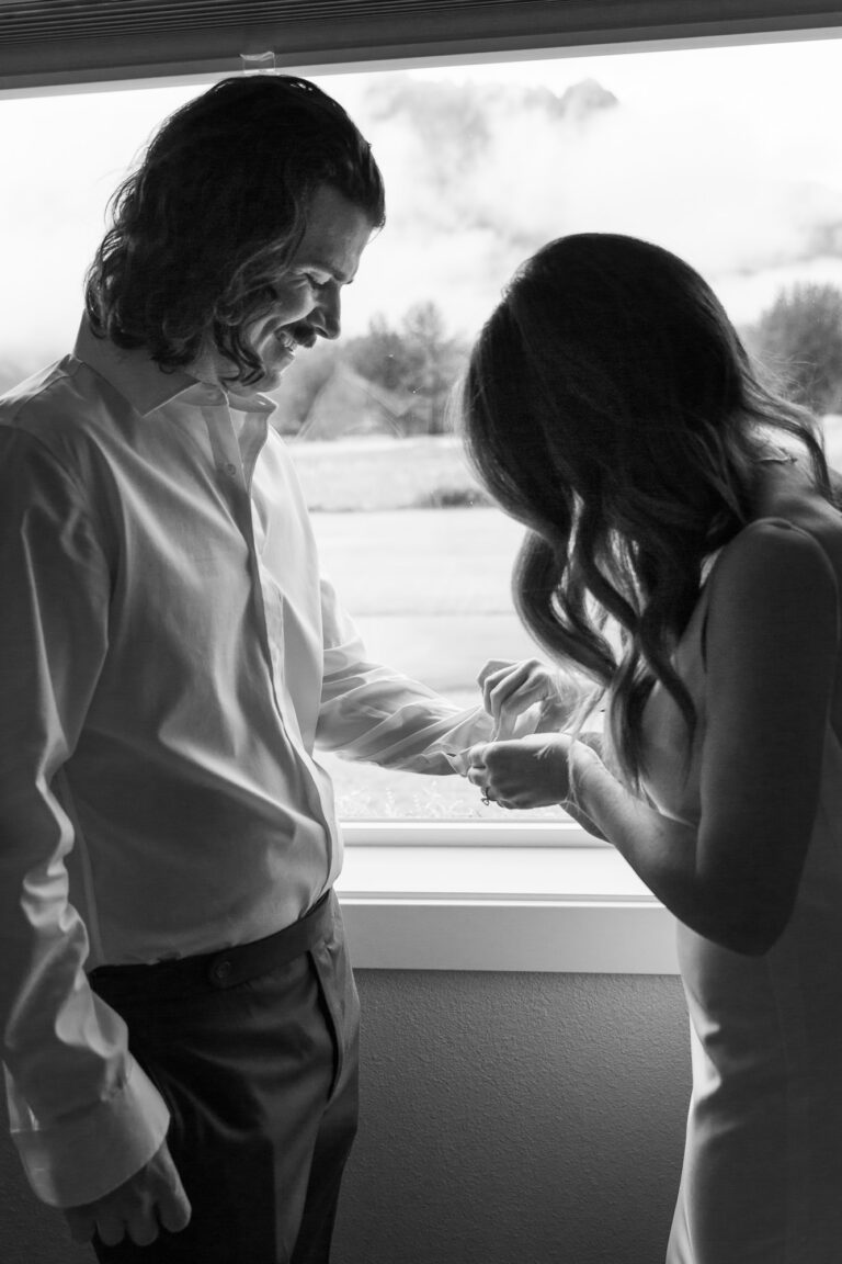 Black and white photo of bride helping the groom fix his cuff links in front of the window at their airbnb in Alaska.