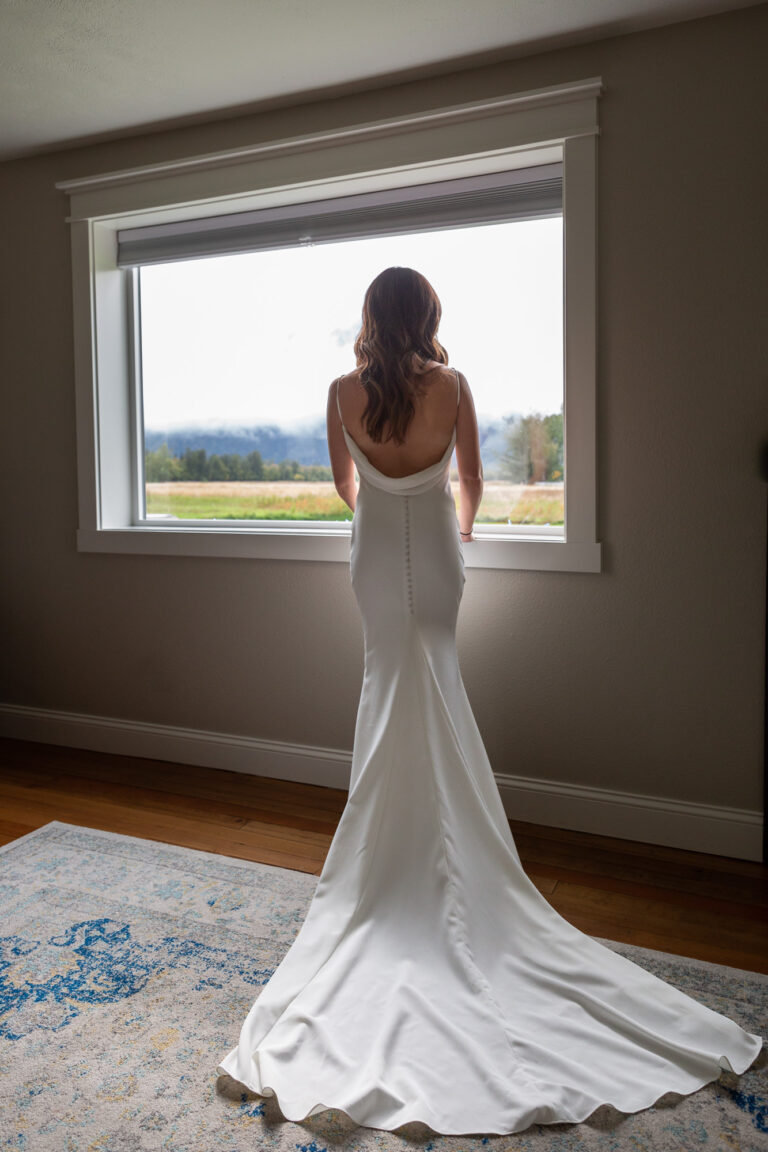 Bride stands looking out the window of the airbnb with her wedding dress laid out behind her on her Alaska Elopement day