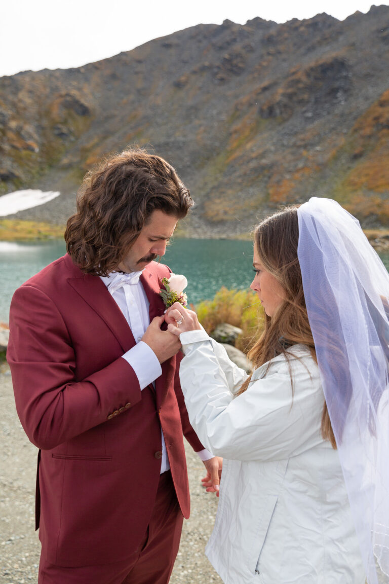 A bride helps pin her groom's boutonniere on his chest during their Hatcher Pass elopement day.