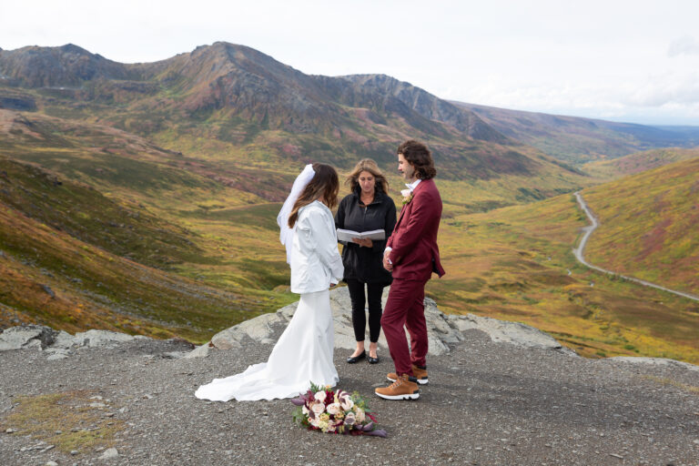 A bride and groom stand facing each other while their Hatcher Pass elopement officiant reads from a book behind them