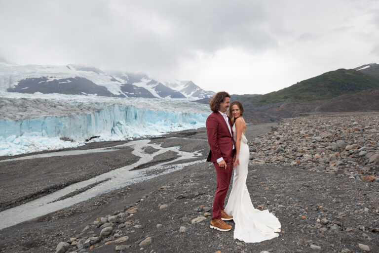 A bride leans against her groom's chest to keep warm while standing in front of a blue glacier on their Hatcher Pass elopement day.