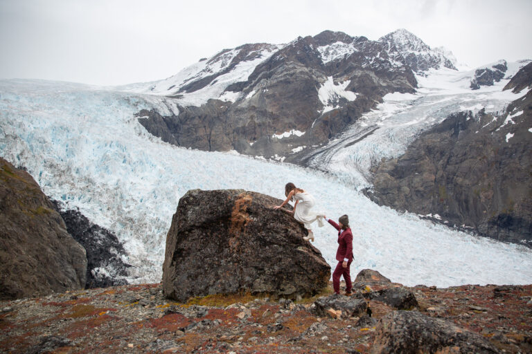 Bride climbs up on a rock while groom holds her wedding dress. There is a glacier field behind them and snow on all of the mountains.