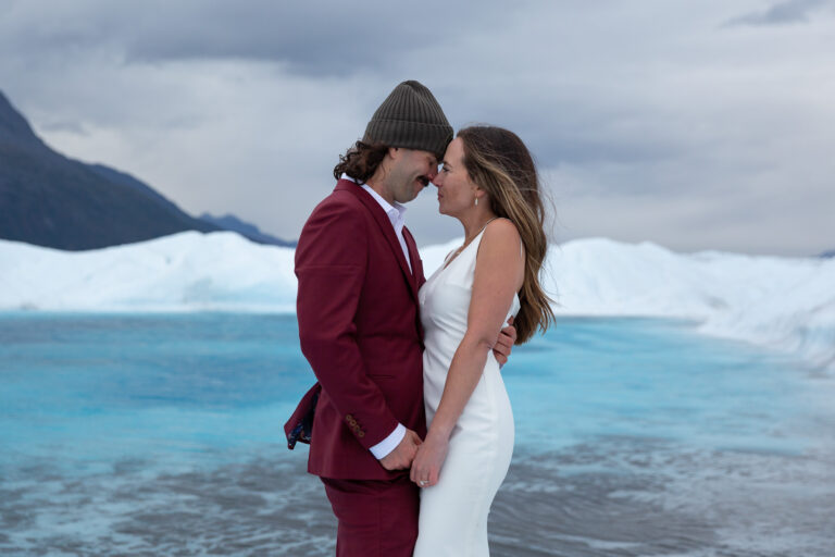 Bride and groom stand facing each other with their foreheads touching in front of a bright blue glacier pool.