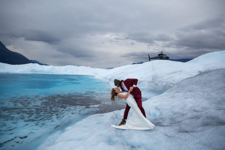 Groom dips bride and kisses her as they stand next to a bright blue glacier pool in Alaska.