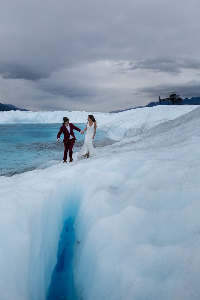 Bride and groom walk on the ice with a glacier pool behind them and a helicopter resting above them on the ice.