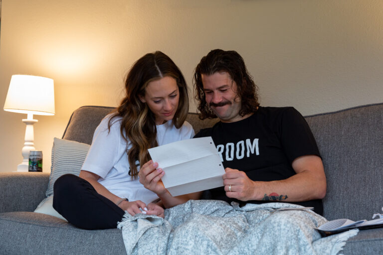 Bride and groom sit next to each other on the couch in their Airbnb reading letters from their family and friends back home.