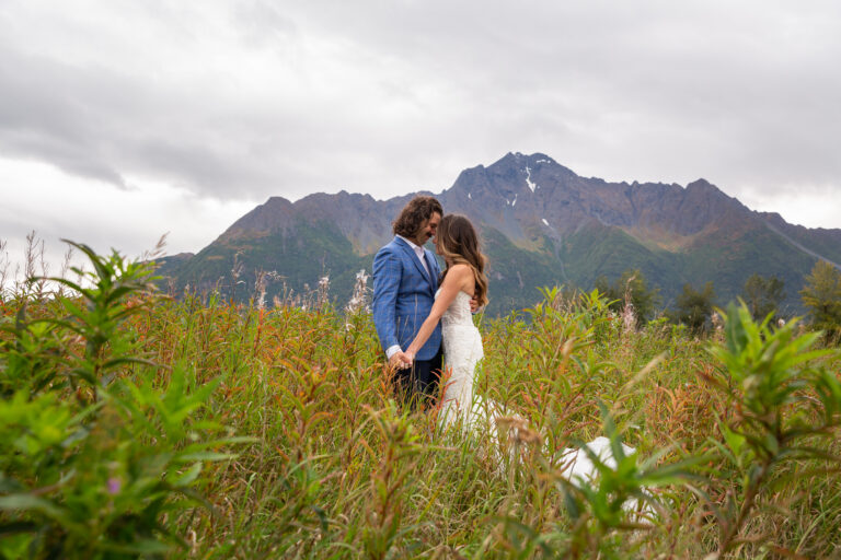 A bride and groom stand with their foreheads touching in a grassy field with flowers around them