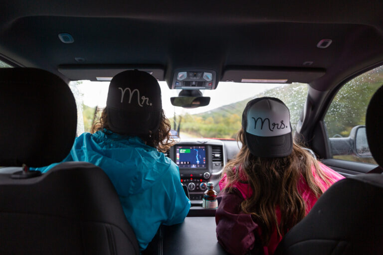 A couple wearing "Mr." and "Mrs." hats backwards, sit in the front seat of a truck.