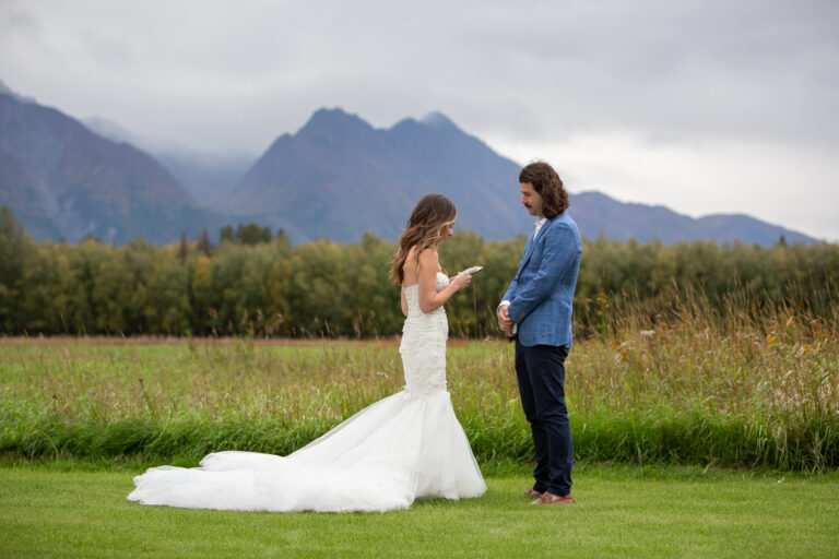 A bride and groom stand in a green field with mountains behind them as the bride reads her vows to her groom.