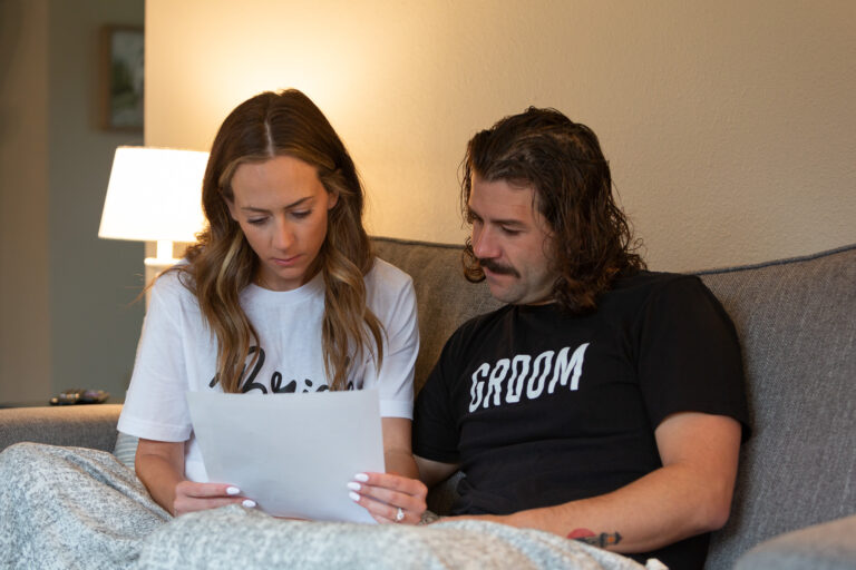 A bride and groom sit on a couch reading a letter together.