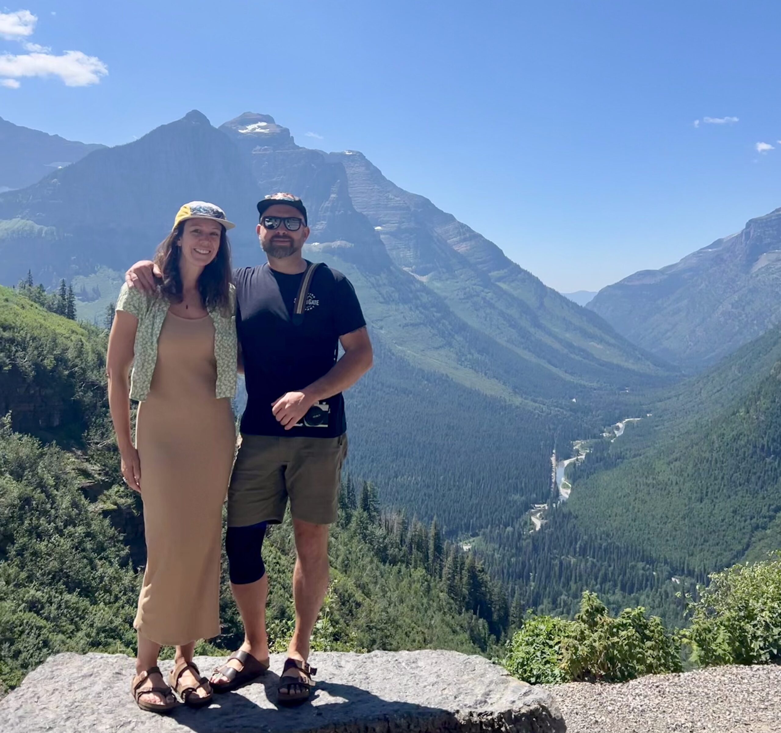 A woman wearing a tan dress and hat stands on a rock in Glacier National Park next to a man in shorts and a t-shirt, with his arm around her shoulders.