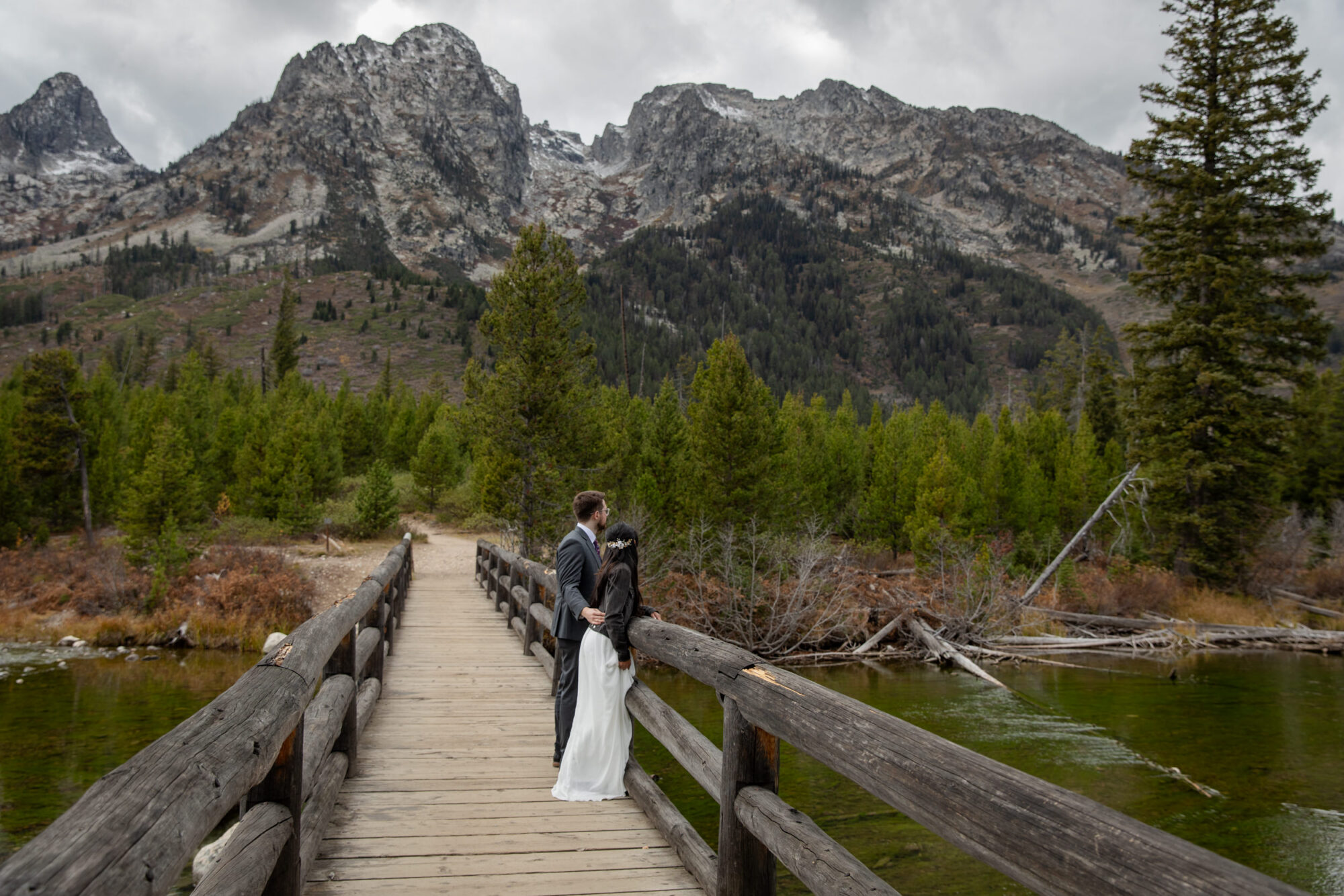 8 Incredible Places to Elope in Wyoming | Scenic Vows Newlyweds standing on a wooden bridge with the Teton mountains behind them in Wyoming