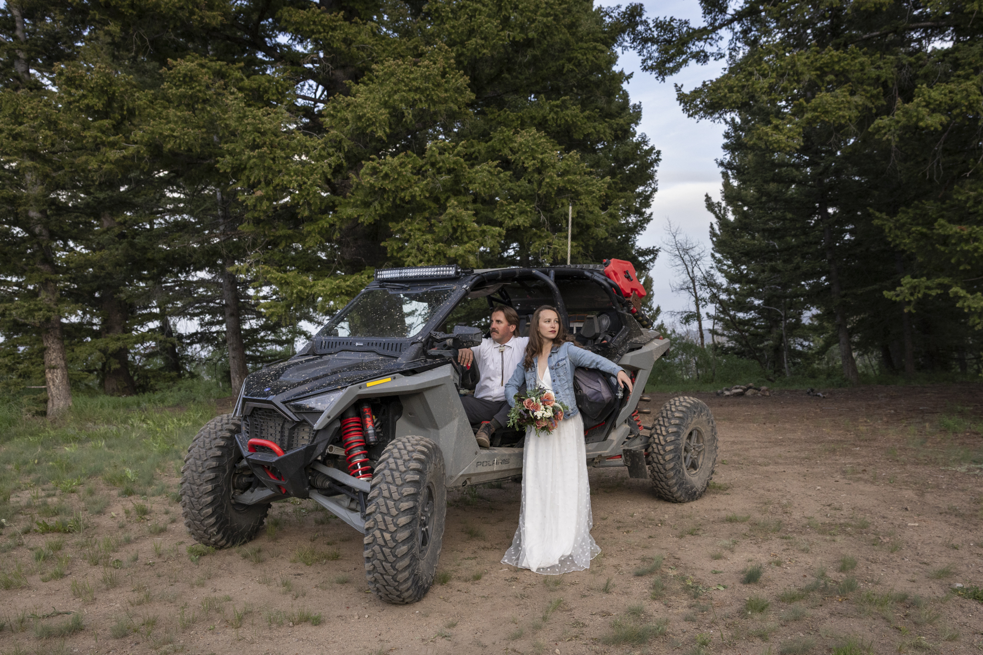 A groom sits in a side-by-side while his bride stands by the front door wearing a white dress and jean jacket and holding a bouquet of flowers on their adventure elopement day.