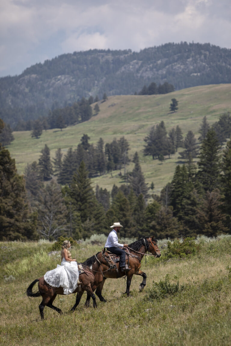 15 Adventure Elopement Ideas: Memorable Ways to Break the Mold | Scenic Vows A bride in a white wedding dress rides a horse up a grassy hill behind her groom who wears a cowboy hat and sits atop his own horse.