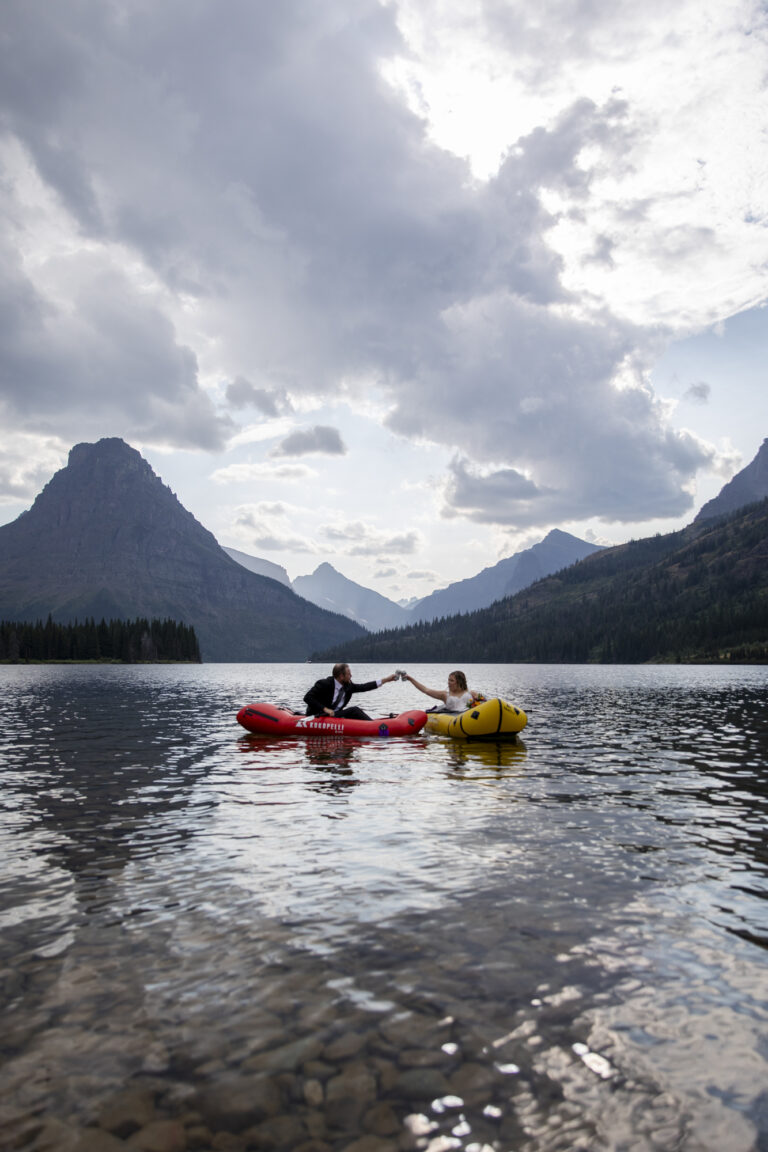 15 Adventure Elopement Ideas: Memorable Ways to Break the Mold | Scenic Vows A bride and groom sit in separate packrafts, one red and one yellow, as they cheers beers in the air over the lake.