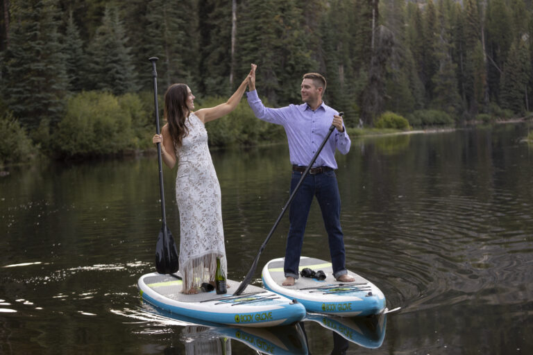 15 Adventure Elopement Ideas: Memorable Ways to Break the Mold | Scenic Vows A bride and groom stand on paddle boards next to each other in a lake in Idaho, high-fiving on their adventure elopement day.