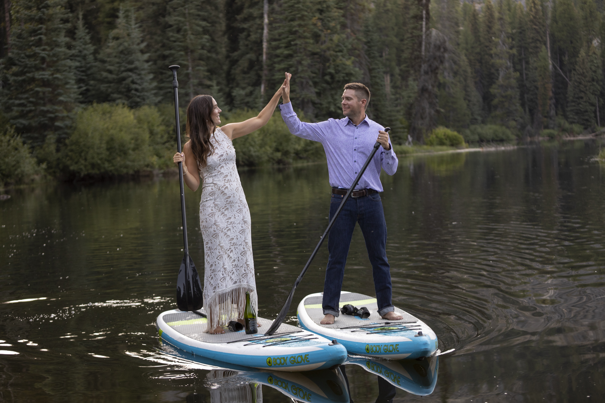 A bride and groom stand on paddle boards next to each other in a lake in Idaho, high-fiving on their adventure elopement day.