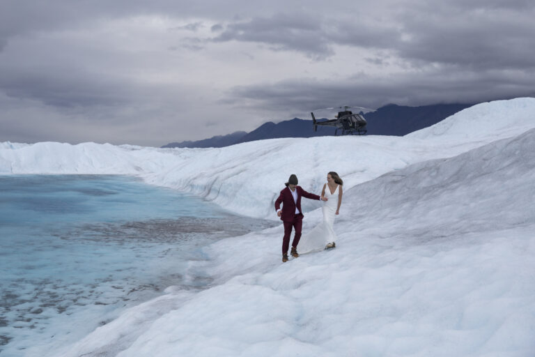 15 Adventure Elopement Ideas: Memorable Ways to Break the Mold | Scenic Vows A bride and groom walk hand in hand along the side of a glacier in Alaska with a helicopter parked in the snow behind them and a vibrant blue glacier lake it to their right.