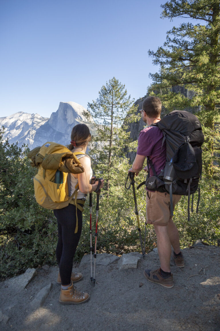 15 Adventure Elopement Ideas: Memorable Ways to Break the Mold | Scenic Vows A man and woman wearing big backpacks and holding trekking poles in their hands, pause on a hike to look out at Half Dome during their Yosemite adventure elopement.