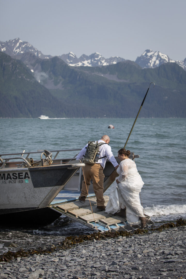 15 Adventure Elopement Ideas: Memorable Ways to Break the Mold | Scenic Vows A groom standing on the front of a fishing boat reaches back to grab the hand of his wife, who is wearing a white wedding dress and brown hiking boots.