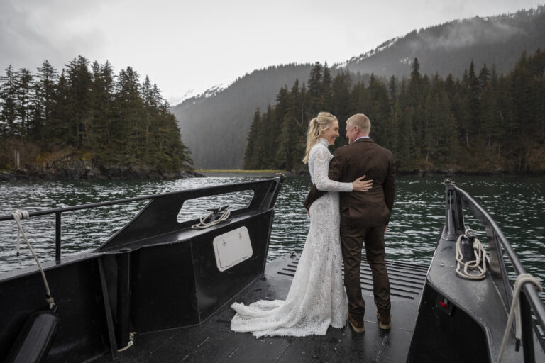 15 Adventure Elopement Ideas: Memorable Ways to Break the Mold | Scenic Vows A bride and groom stand with their arms around each others waist, at the edge of a boat smiling at each other as the boat heads to land on a forested island in alaska.