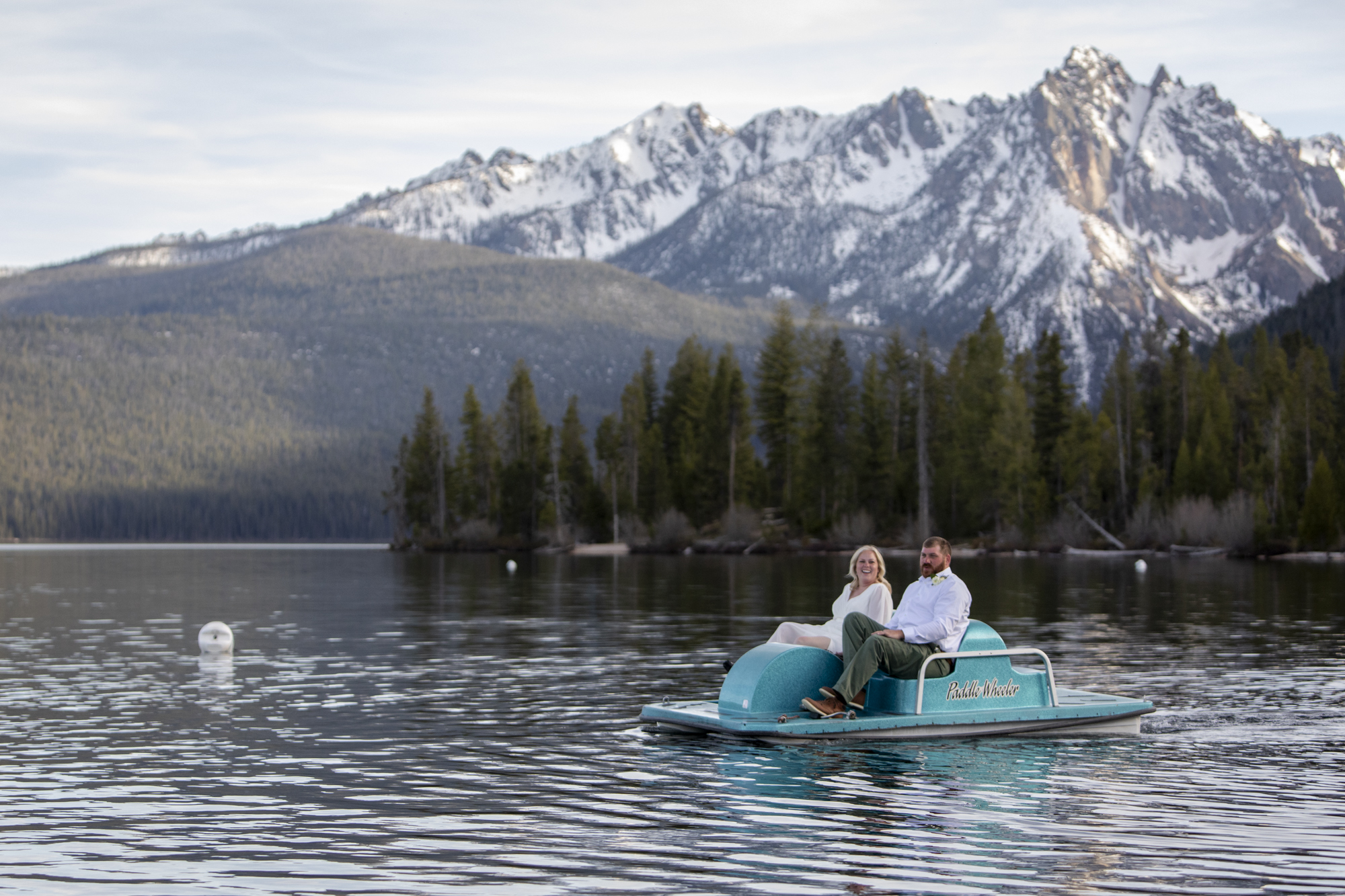 A man wearing green pants and a white shirt sits next to a woman wearing a wedding dress in a paddle boat in the middle of a lake with snowy mountains and a forest behind them.