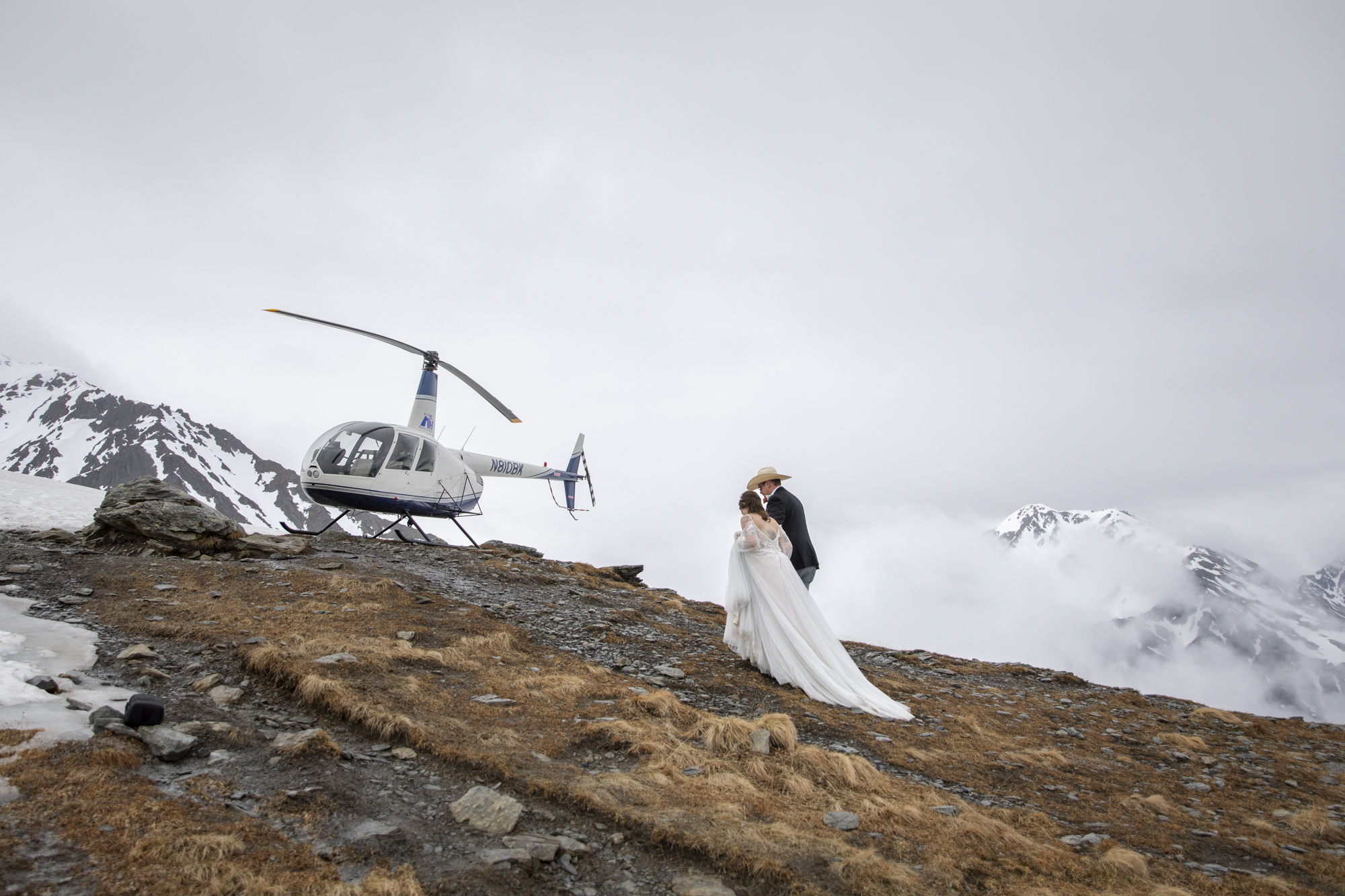 A bride wearing a long white wedding dress walks up the side of a grassy hill towards a parked helicopter next to a man wearing a cowboy hat.