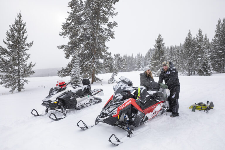 15 Adventure Elopement Ideas: Memorable Ways to Break the Mold | Scenic Vows A bride and groom stand in the snow packing a bag on the back of a snowmobile, laughing and smiling at each other on their elopement day.