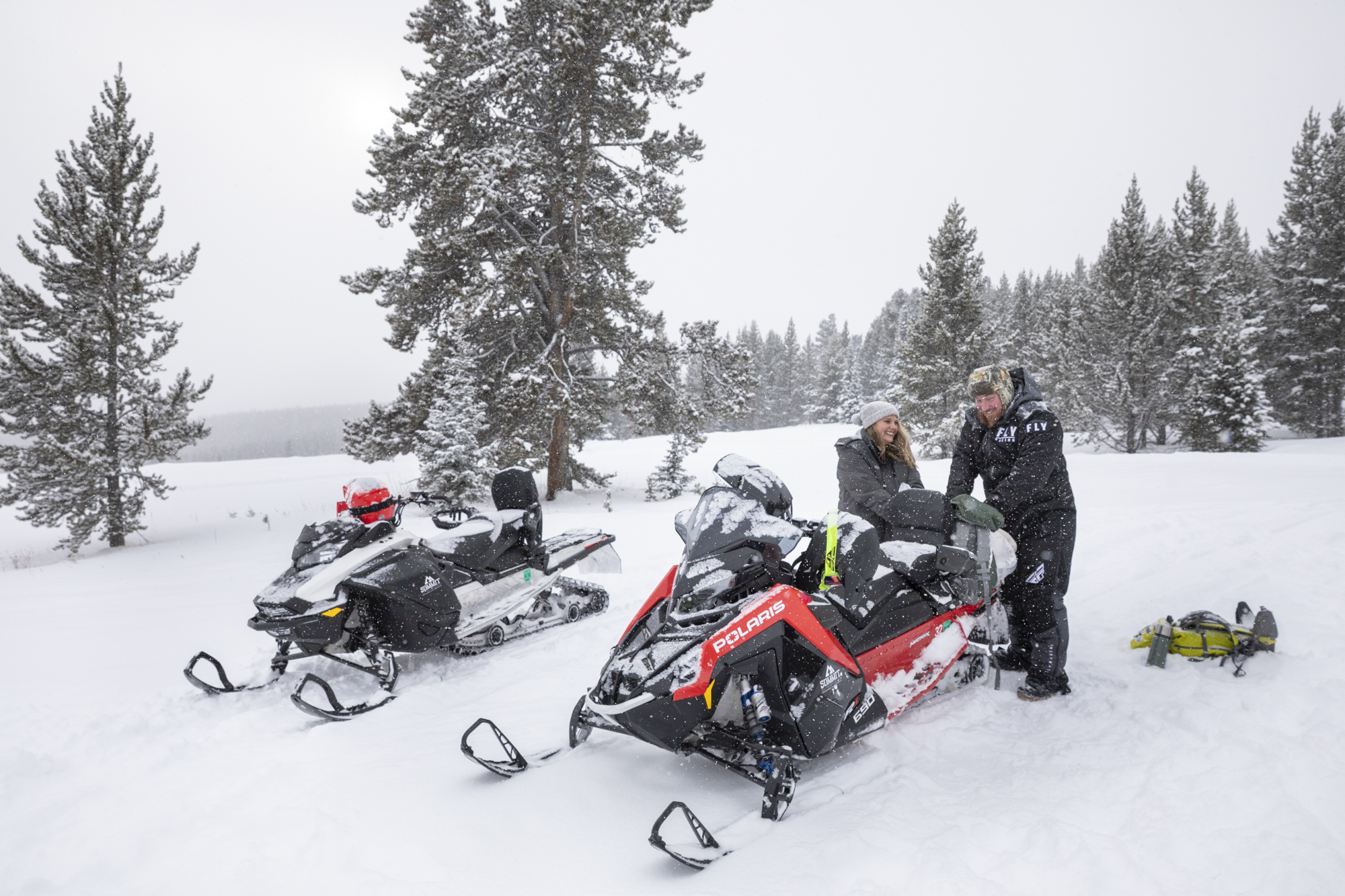 Eloping in Glacier National Park: Your Complete Guide | Scenic Vows A bride and groom stand in the snow packing a bag on the back of a snowmobile, laughing and smiling at each other after eloping in Glacier National Park.