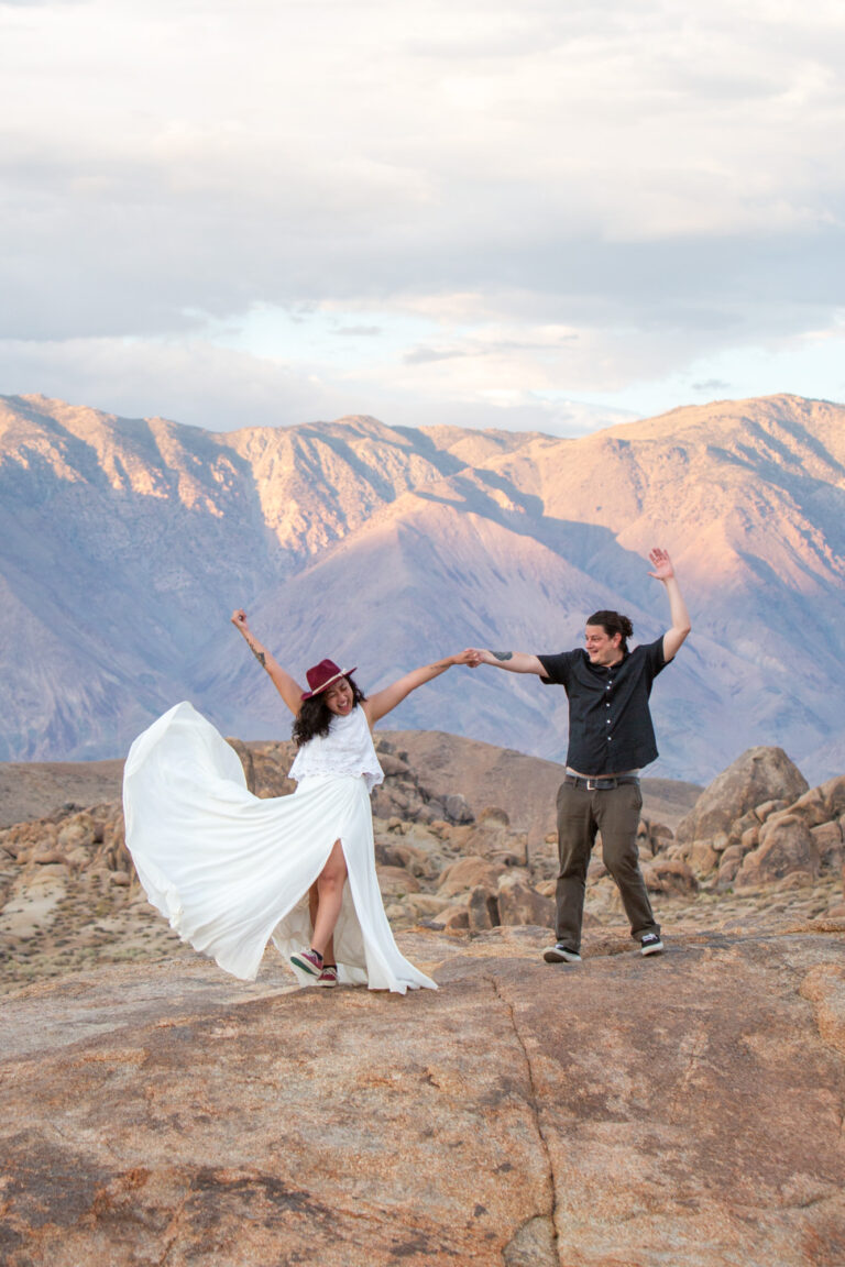 A bride and groom throw their hands up in the air and the bride kicks her dress up in celebration on their Alabama Hills adventure elopement day.