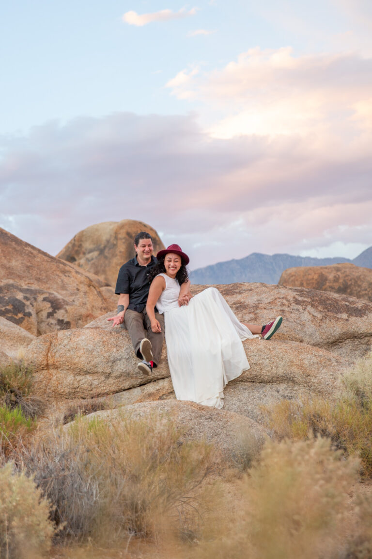 A bride and groom smile and laugh while sitting next to each other on a rock in Alabama Hills.
