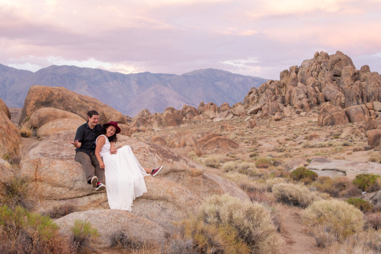 A bride and groom sit on a rock laughing as the sun sets behind them in the Alabama Hills