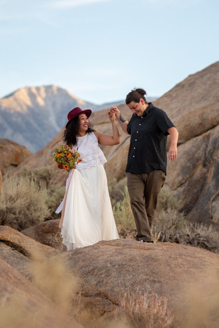A bride and groom hold hands as they walk across big boulders in Alabama Hills.