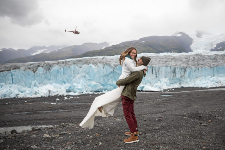 A groom holds his bride in the air as the stand in front of a glacier and a helicopter flies overhead.