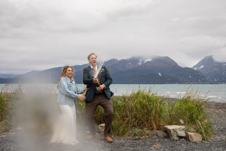 A groom stands on a rocky beach in Alaska, spraying a champagne bottle as his bride stands next to him looking shocked and laughing.