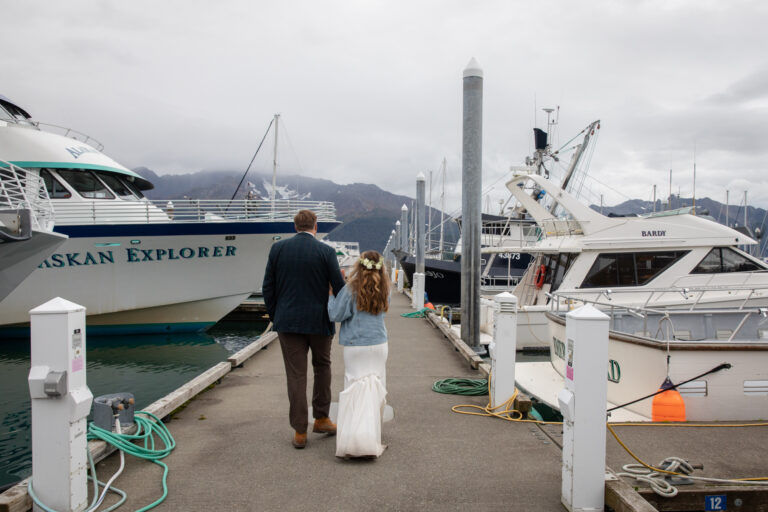 A bride and groom walk arm in arm down a path in a harbor in Alaska with boats on either side of them.