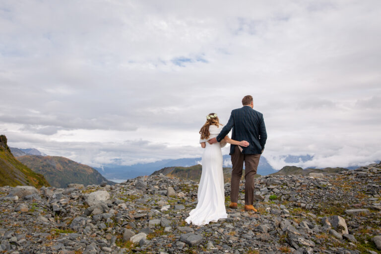 A couple stand with their arms around each other's back as the take in the beautiful mountain views on their Alaska elopement day.
