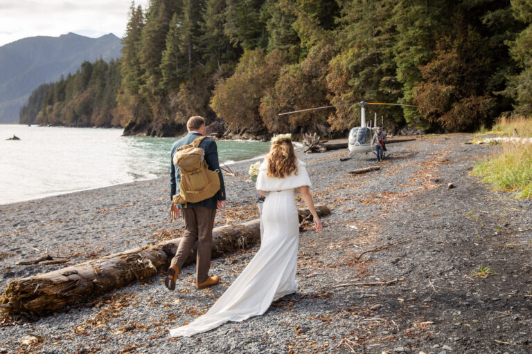 A bride and groom walk along a rocky beach towards a helicopter that is parked waiting to take them away for their Alaska elopement ceremony.