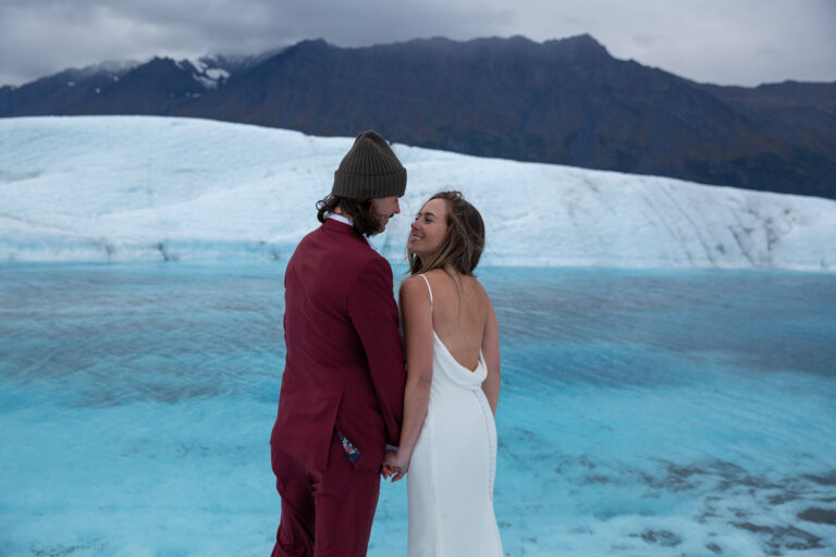 A bride and groom stand next to each other holding hands and smiling in front of a blue glacier pool on their Alaska elopement day.